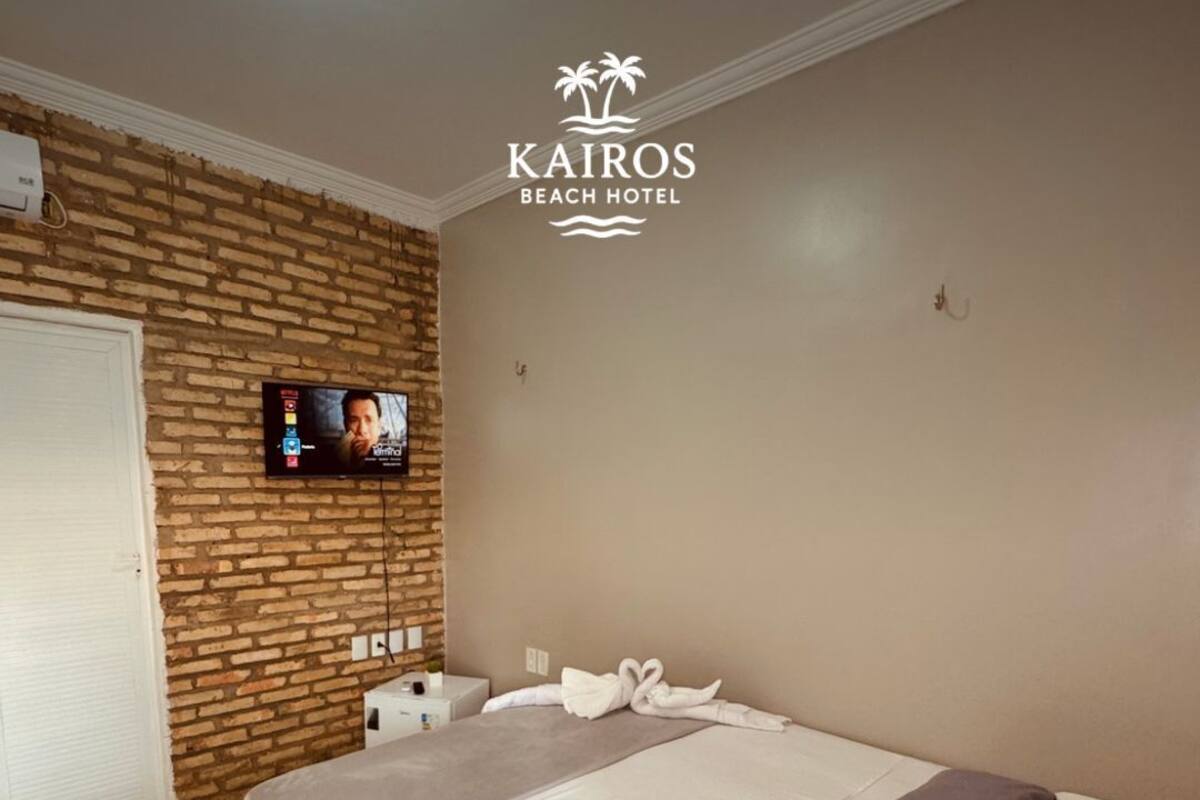 Habitación de hotel con pared de ladrillo, un televisor montado en la pared, una cama con ropa de cama blanca y marrón, y el logotipo de KAIROS BEACH HOTEL en la pared.