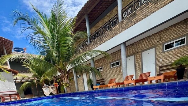 Une piscine à l’eau d’un bleu cristallin, entourée d’un bâtiment à la façade en briques et dotée d’un balcon avec des chaises en bois.