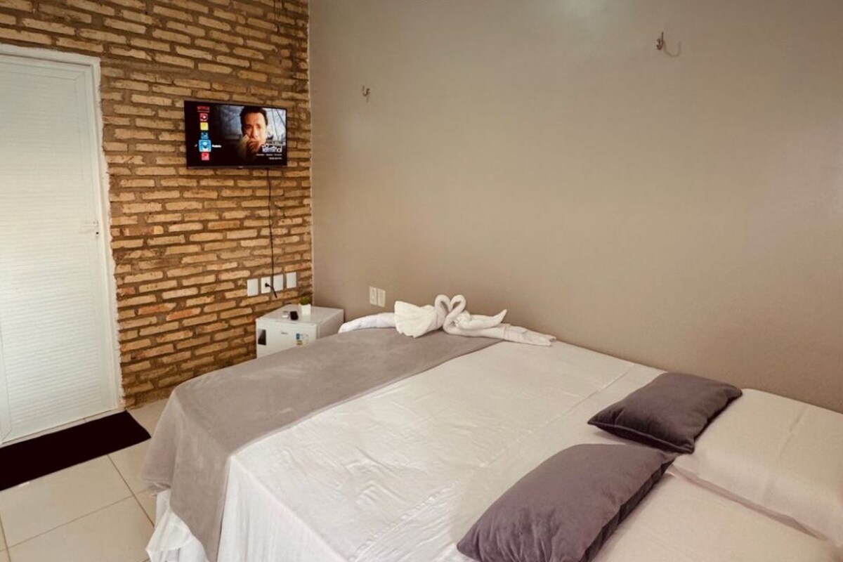 Quarto de hotel com parede de tijolos, televisão fixada na parede, cama com roupa de cama branca e marrom, e o logotipo do KAIROS BEACH HOTEL na parede.