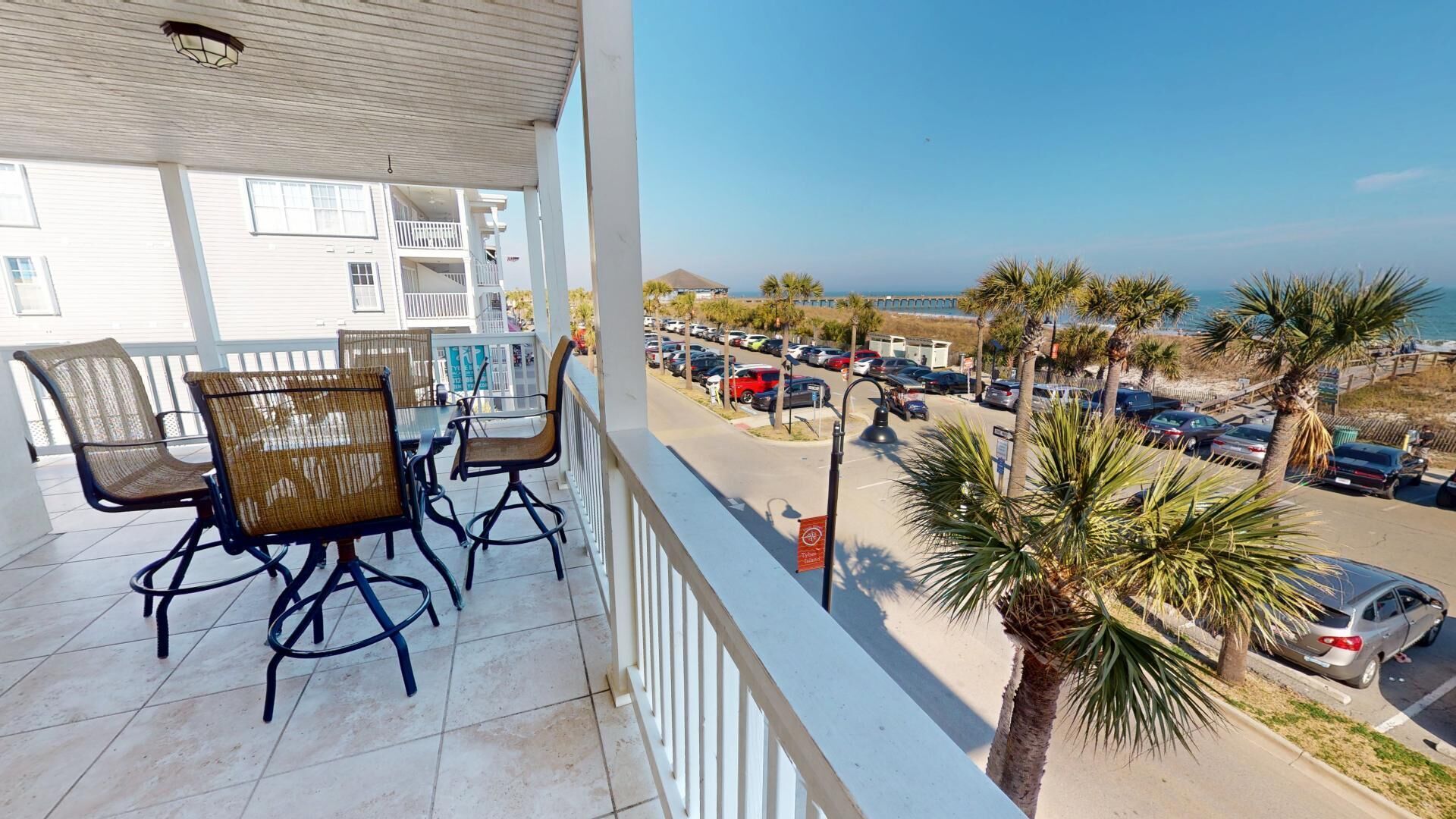En balkon med fletstole og et bord, der vender ud mod en parkeringsplads og en strand.