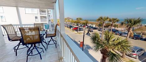 En balkon med fletstole og et bord, der vender ud mod en parkeringsplads og en strand.