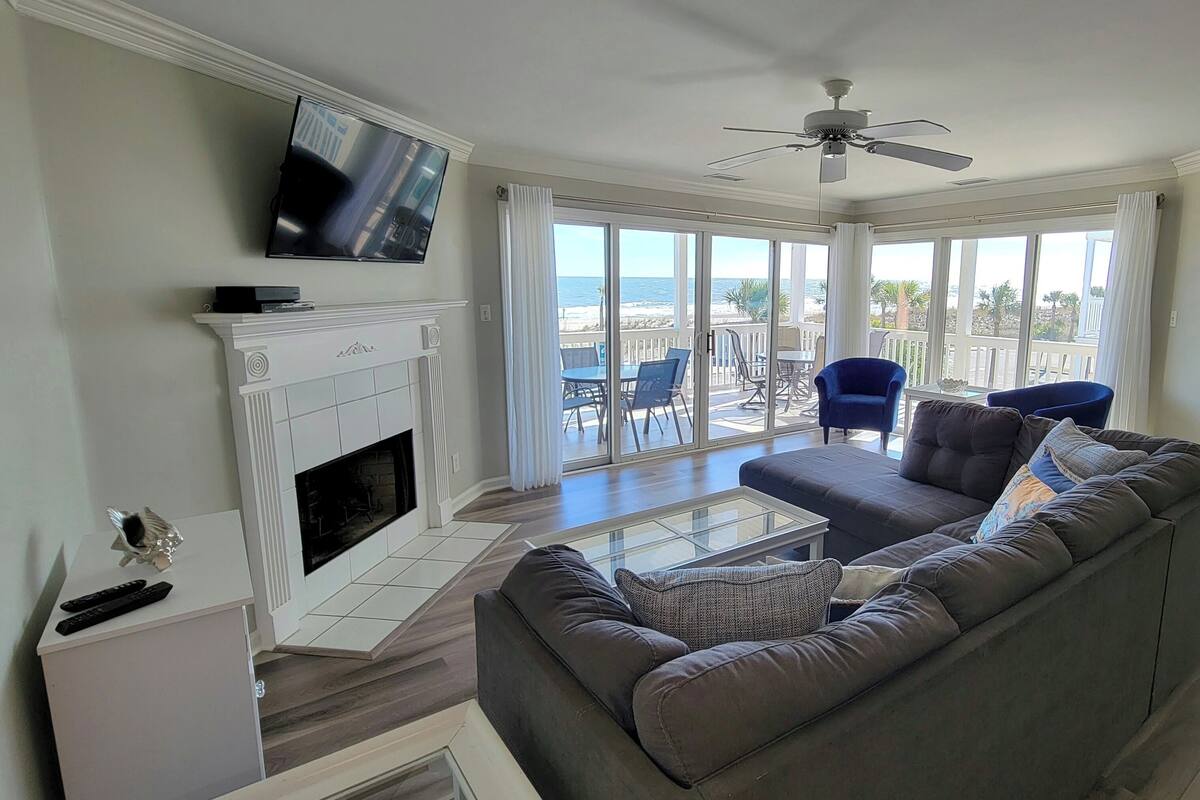A living room with a fireplace, a ceiling fan, a sofa, and a view of the ocean through large windows.