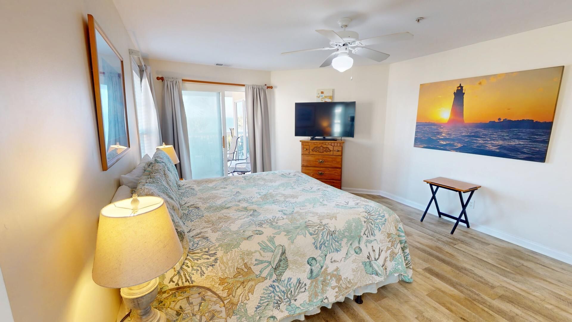 Un dormitorio con cama, televisión, un cuadro de un atardecer y ventilador de techo.