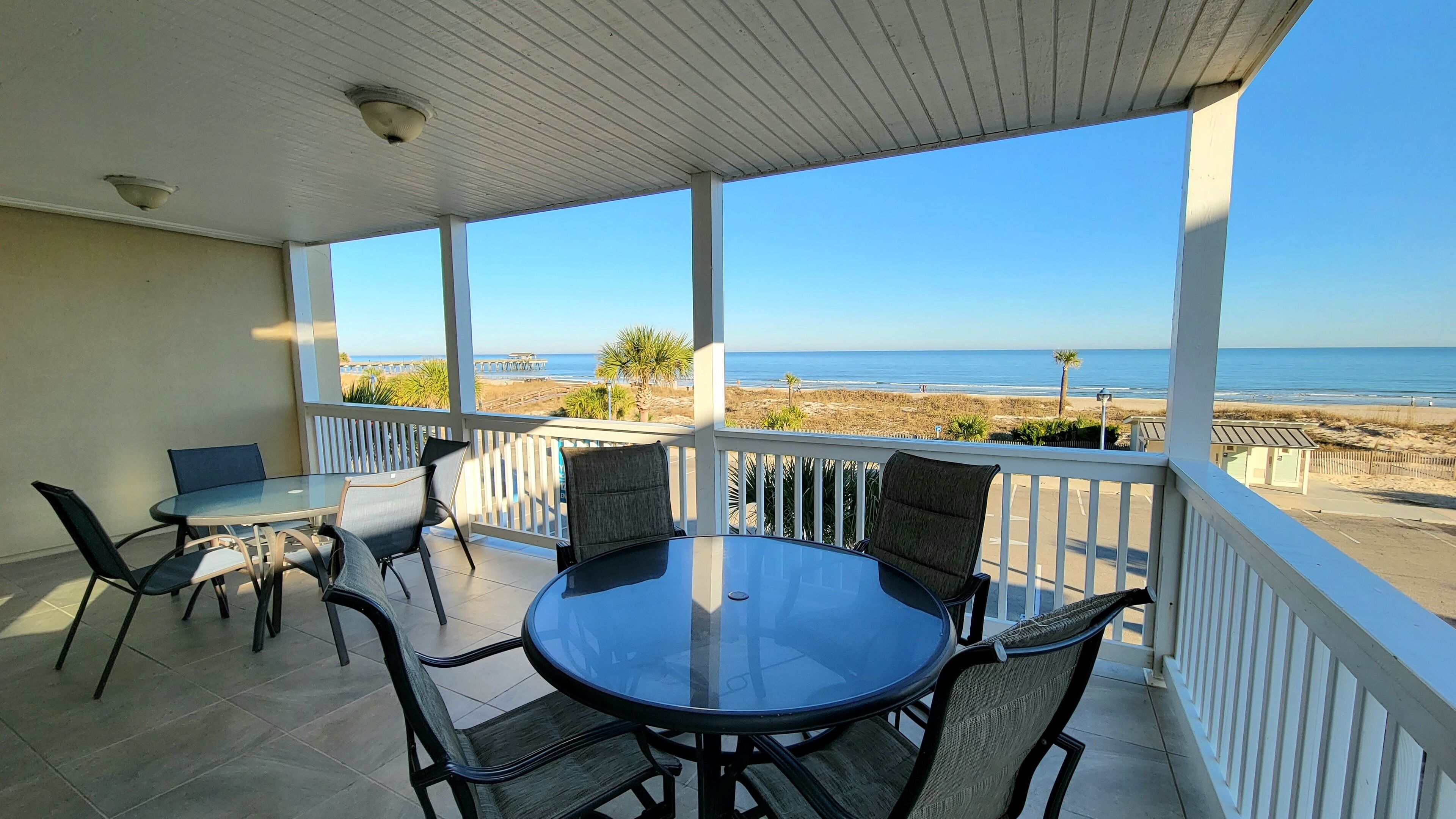 A covered patio with a glass table and chairs, overlooking a beach with a pier and palm trees.