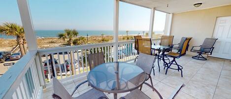 A covered patio with a glass table and chairs, overlooking a beach.