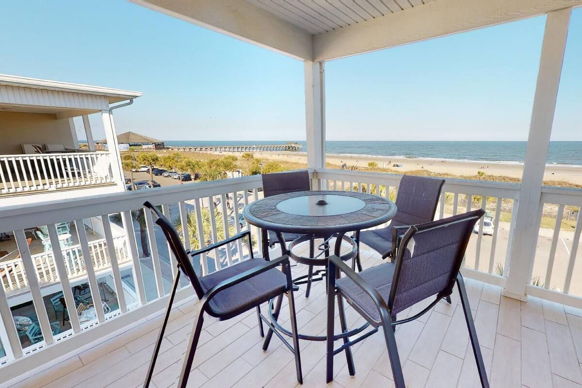 A balcony with a round table and four chairs, overlooking a beach with a clear view of the ocean.