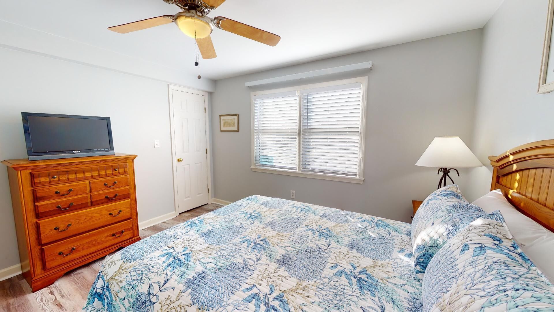 A bedroom with a bed, a dresser, a ceiling fan, a window with blinds, and a TV on the dresser.