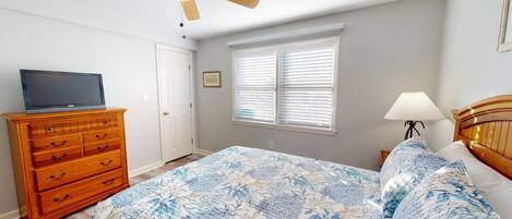 A bedroom with a bed, a dresser, a ceiling fan, a window with blinds, and a TV on the dresser.