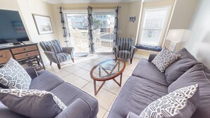 A living room with a grey sofa set, patterned cushions, a glass coffee table, and a TV on a wooden stand. - Gull Reef 614 Tybee Vacation Rentals (Tybee Island)