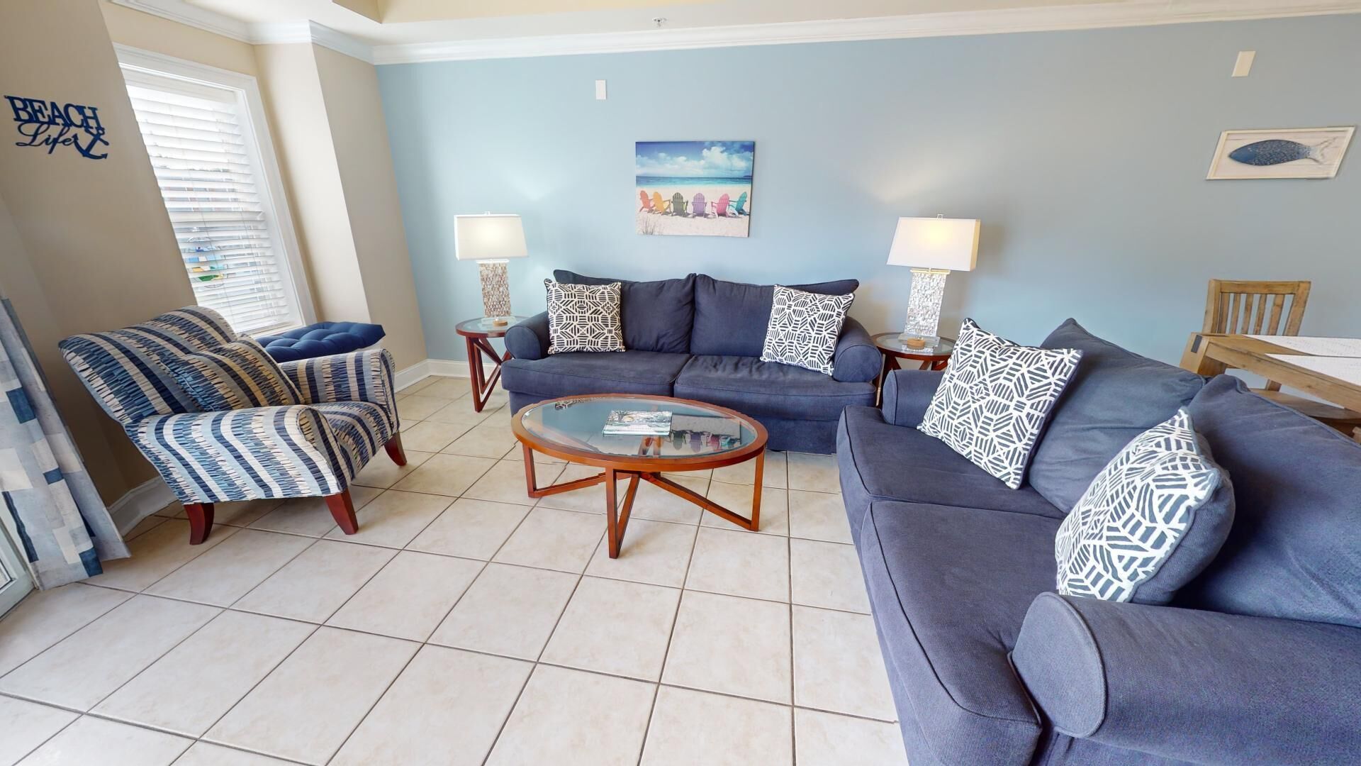 A living room with a blue sofa, a patterned armchair, a glass coffee table, and two side tables with lamps.