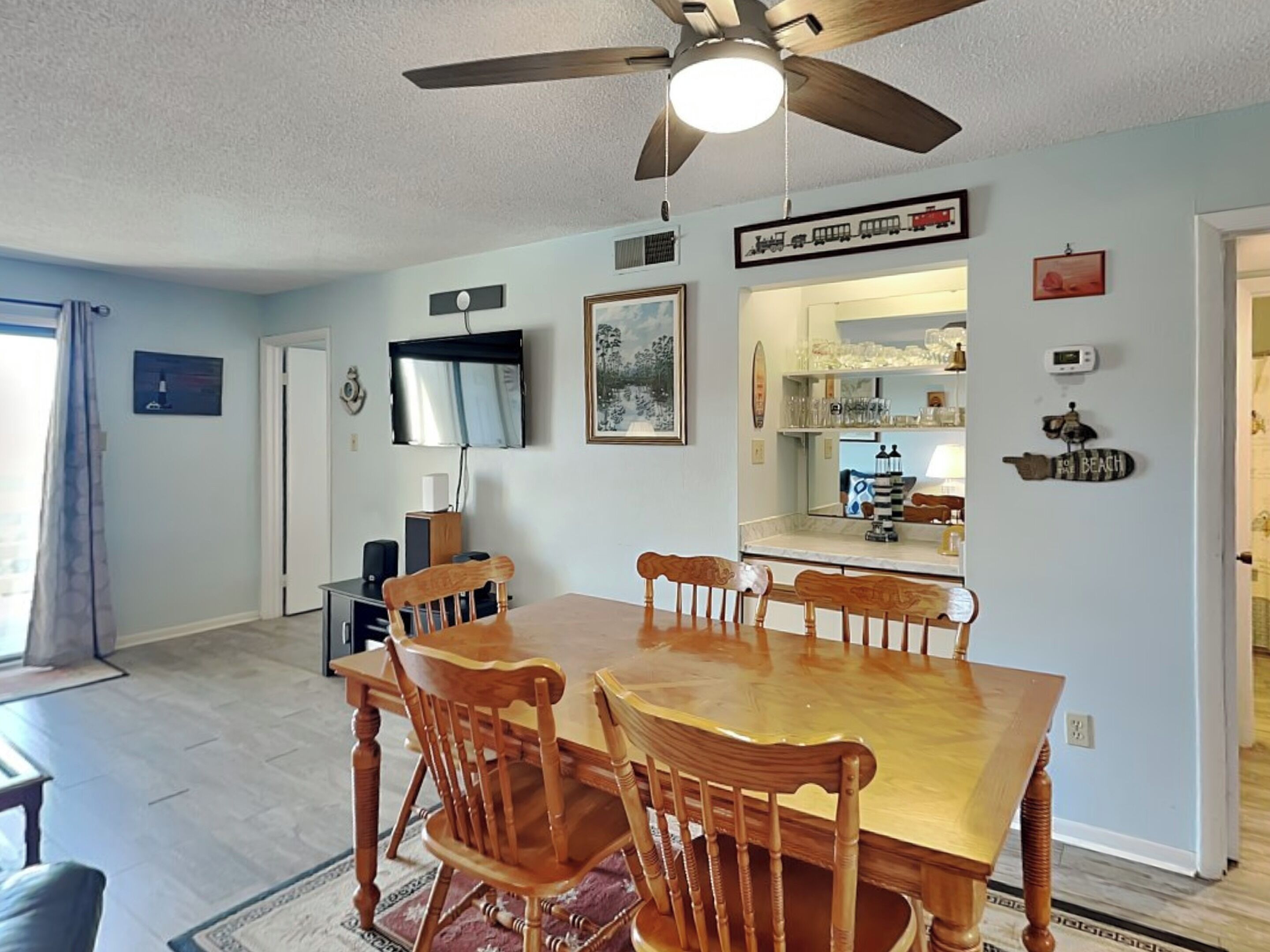 A dining area with a wooden table and chairs, a ceiling fan, a TV mounted on the wall, and a kitchen area with a refrigerator.