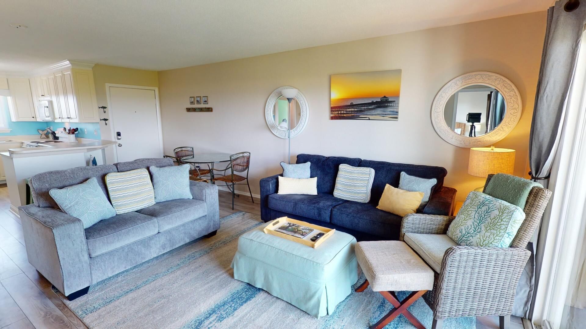 A living room with a grey sofa, a blue sofa, a wicker chair, and a small table with a book. There is a painting on the wall and a mirror above the blue sofa.