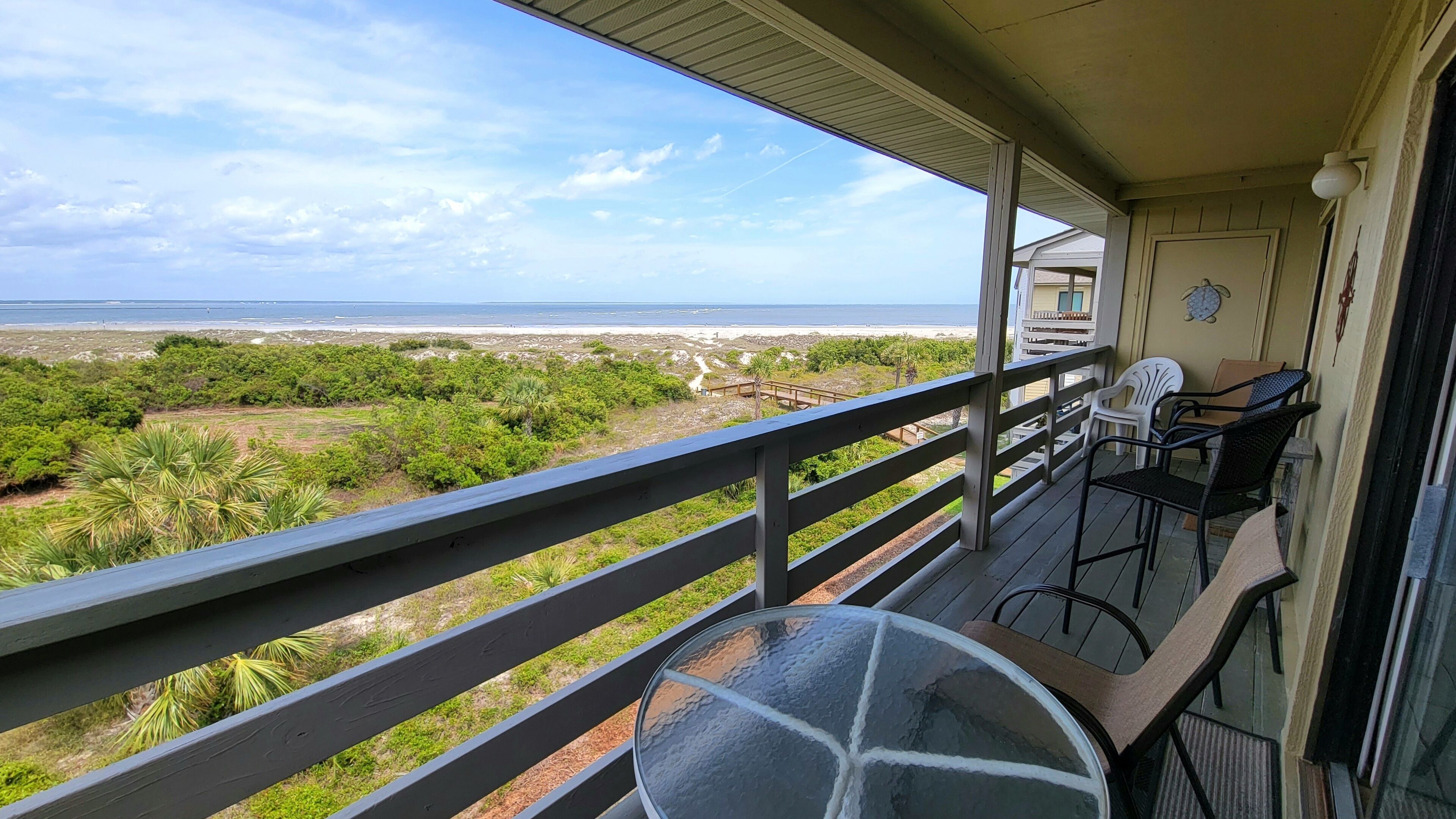 A balcony with a glass table and chairs, overlooking a beach and greenery.