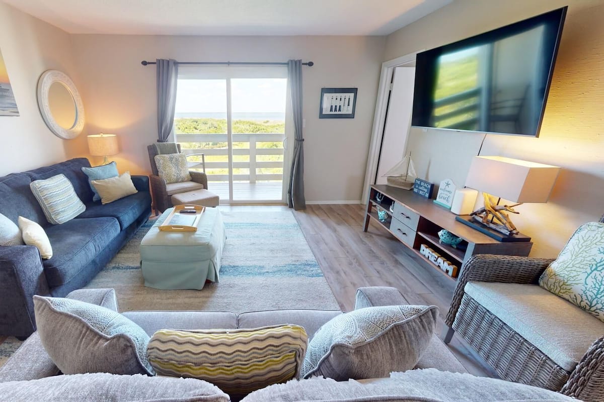 Un salon avec un canapé bleu foncé, un pouf bleu clair et un canapé gris. Un téléviseur est fixé au mur, il y a une fenêtre avec des rideaux et une vue sur l’extérieur.
