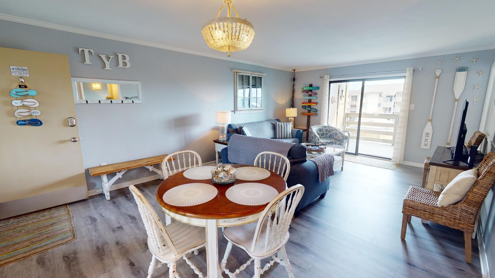 A cozy living room with a dining area, featuring a chandelier, a large window, and a wall with a 'TYB' sign.