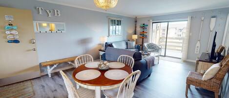 A cozy living room with a dining area, featuring a chandelier, a large window, and a wall with a 'TYB' sign.