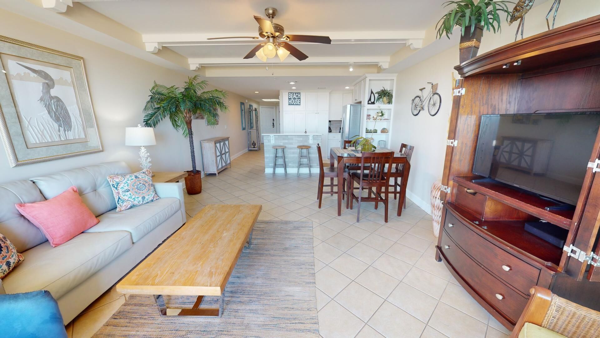 A spacious living area with a sofa, coffee table, and TV stand. There is a dining area with a table and chairs, and a kitchen in the background.