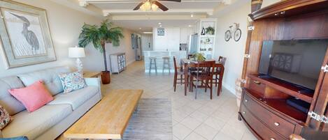 A spacious living area with a sofa, coffee table, and TV stand. There is a dining area with a table and chairs, and a kitchen in the background.