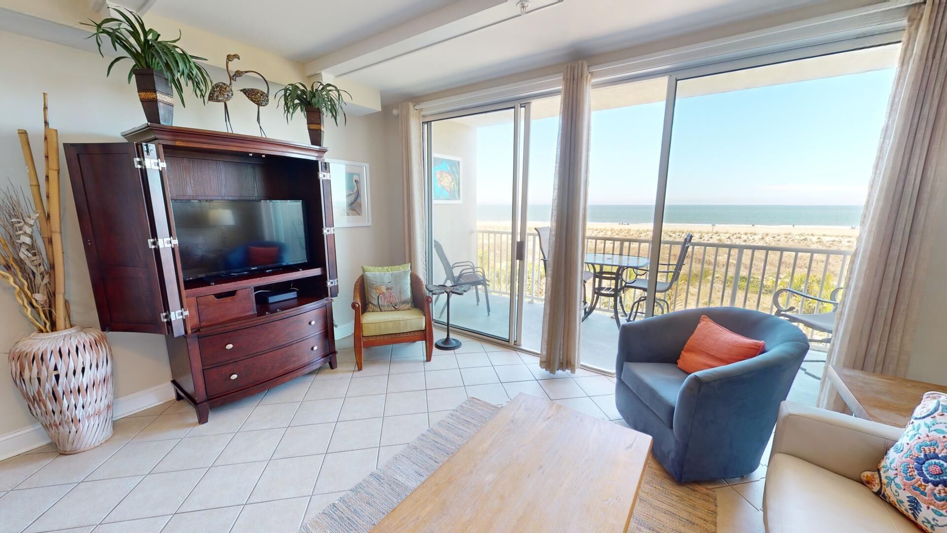 A living room with a TV, sofa, and a balcony with a view of the beach.