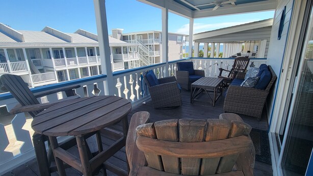 A covered outdoor area with wooden furniture, including a table and chairs, and a wicker sofa with cushions. - Oceanview A by Tybee Vacation Rentals (Tybee Island)