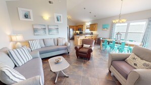 A spacious living room with a sofa, armchairs, and a coffee table. A kitchen with wooden cabinets and a dining area with turquoise chairs are visible in the background. - Oceanview A by Tybee Vacation Rentals (Tybee Island)
