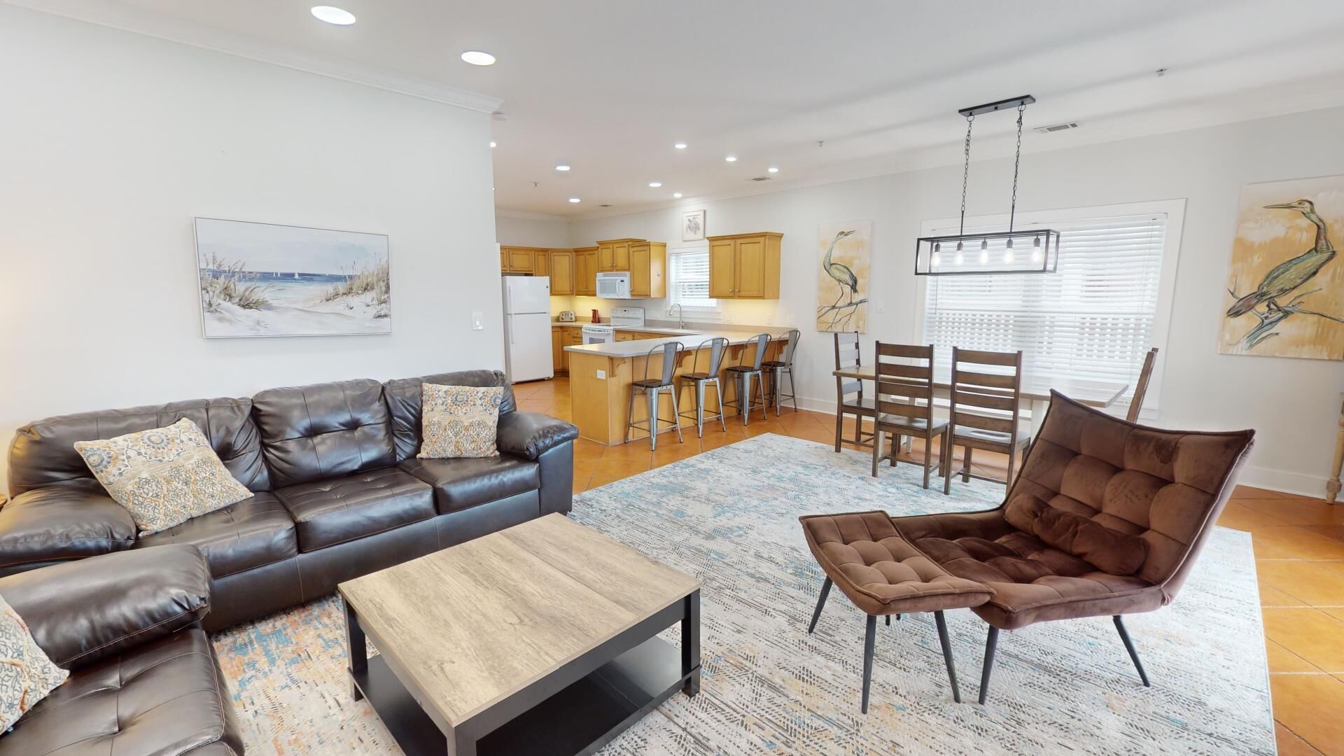 A modern living room with a leather sofa, a wooden coffee table, and a brown armchair. There is a kitchen in the background with wooden cabinets and a dining area with a table and chairs.