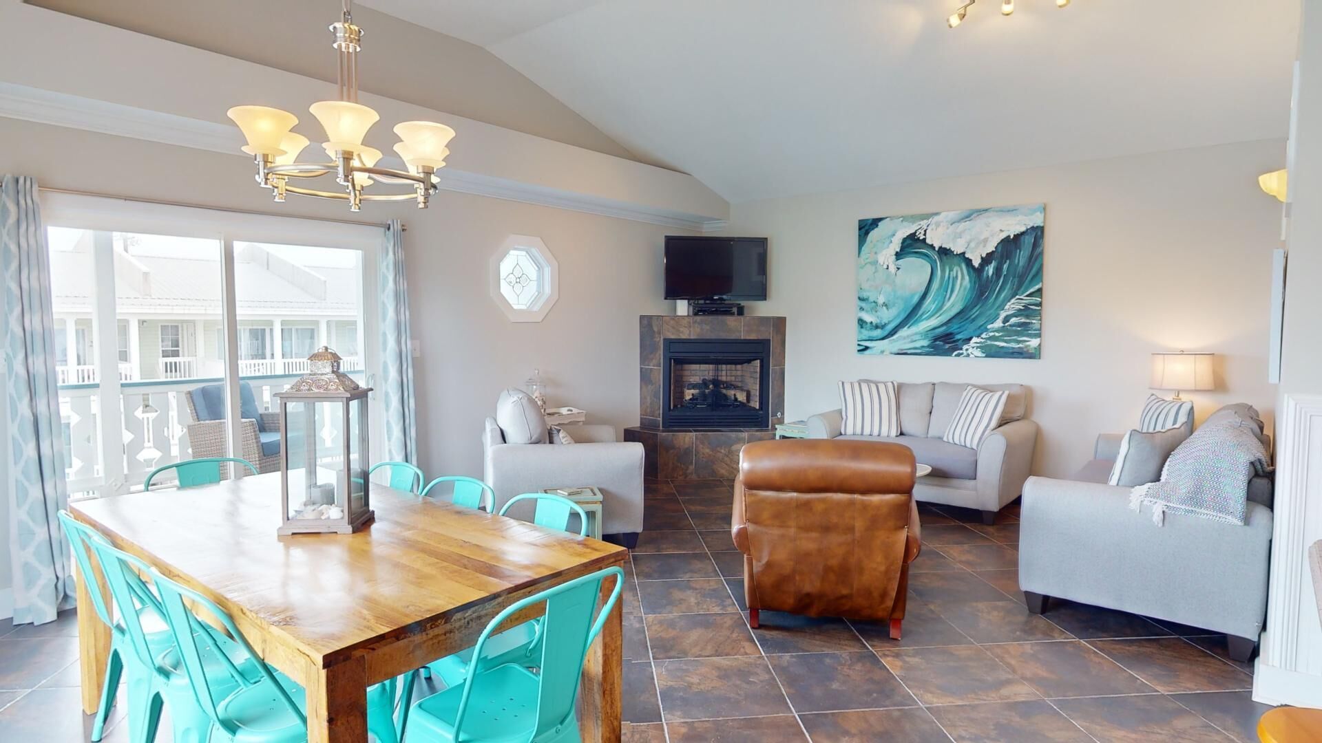 A spacious living area with a wooden dining table, teal chairs, a leather ottoman, and a grey sofa. A fireplace and a large wave painting adorn the walls.