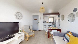 A modern living room with a flat-screen TV, a comfortable sofa, a dining area with a table and chairs, a kitchen in the background, and a wall clock.