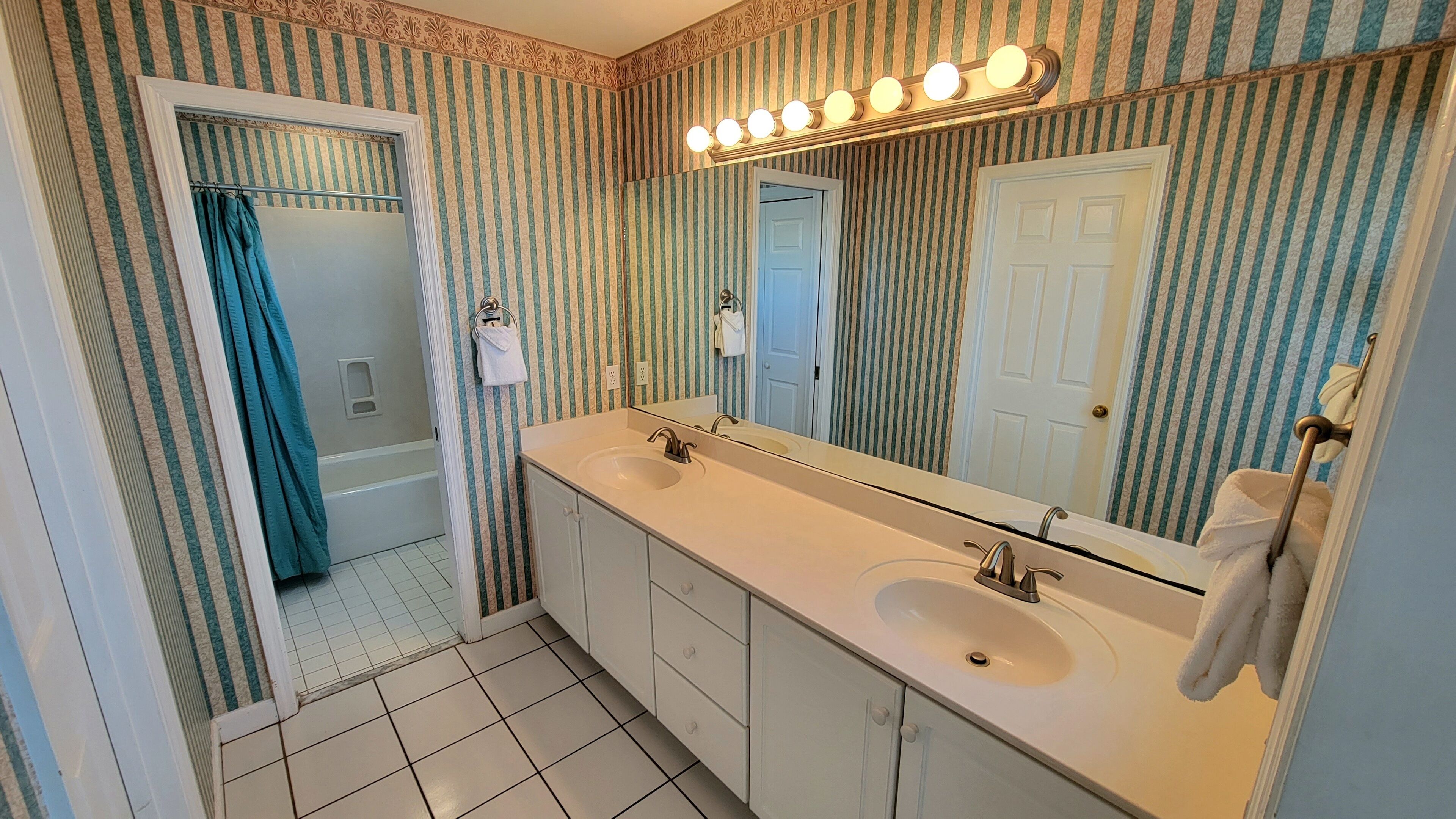 A bathroom with a double sink, a large mirror with lights, striped wallpaper, and a bathtub.