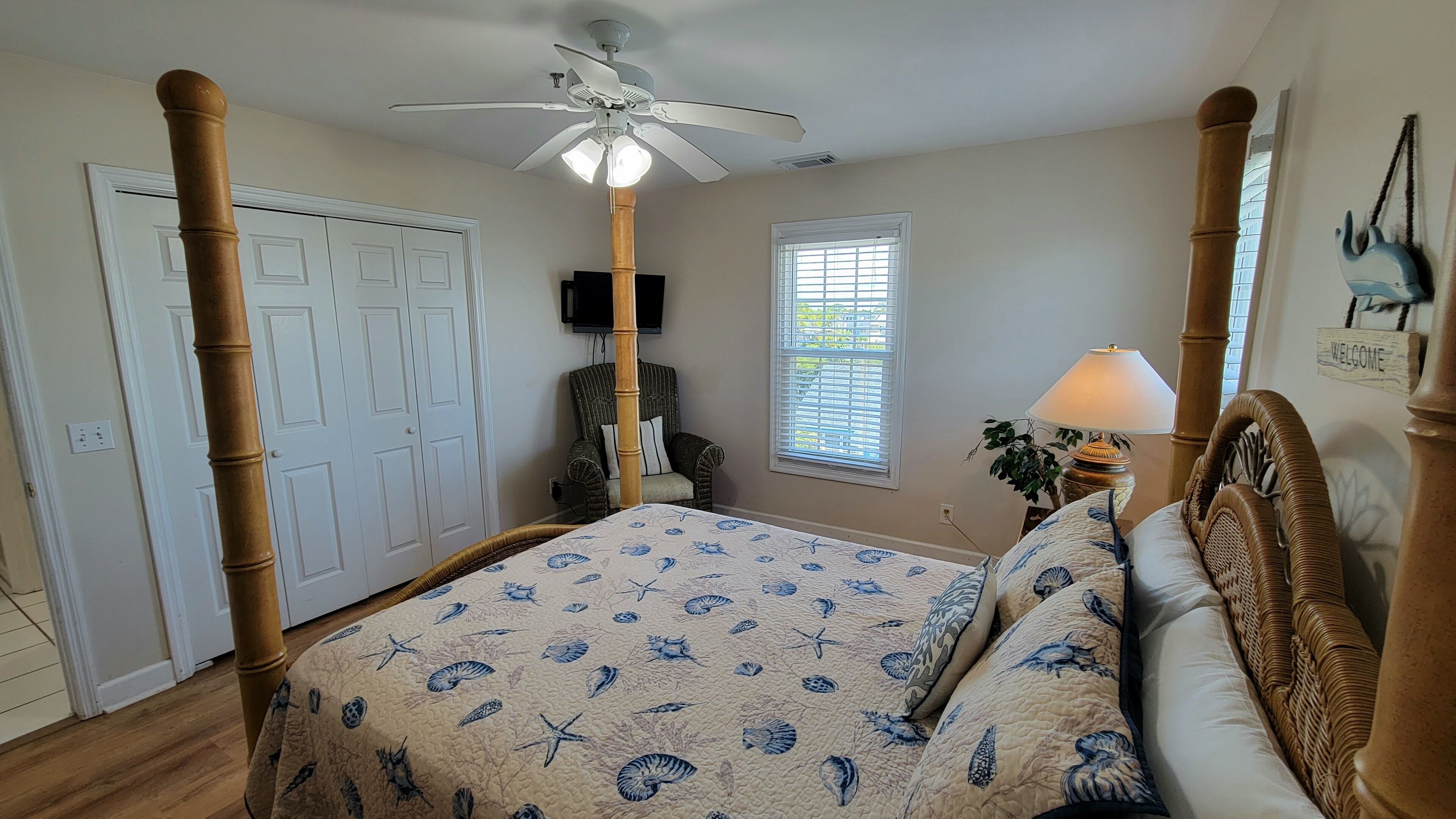 A bedroom with a four-poster bed, a ceiling fan, a window with blinds, a lamp, and a 'Welcome' sign.