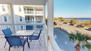 Un balcon avec une table et des chaises, donnant sur une plage et un parking.