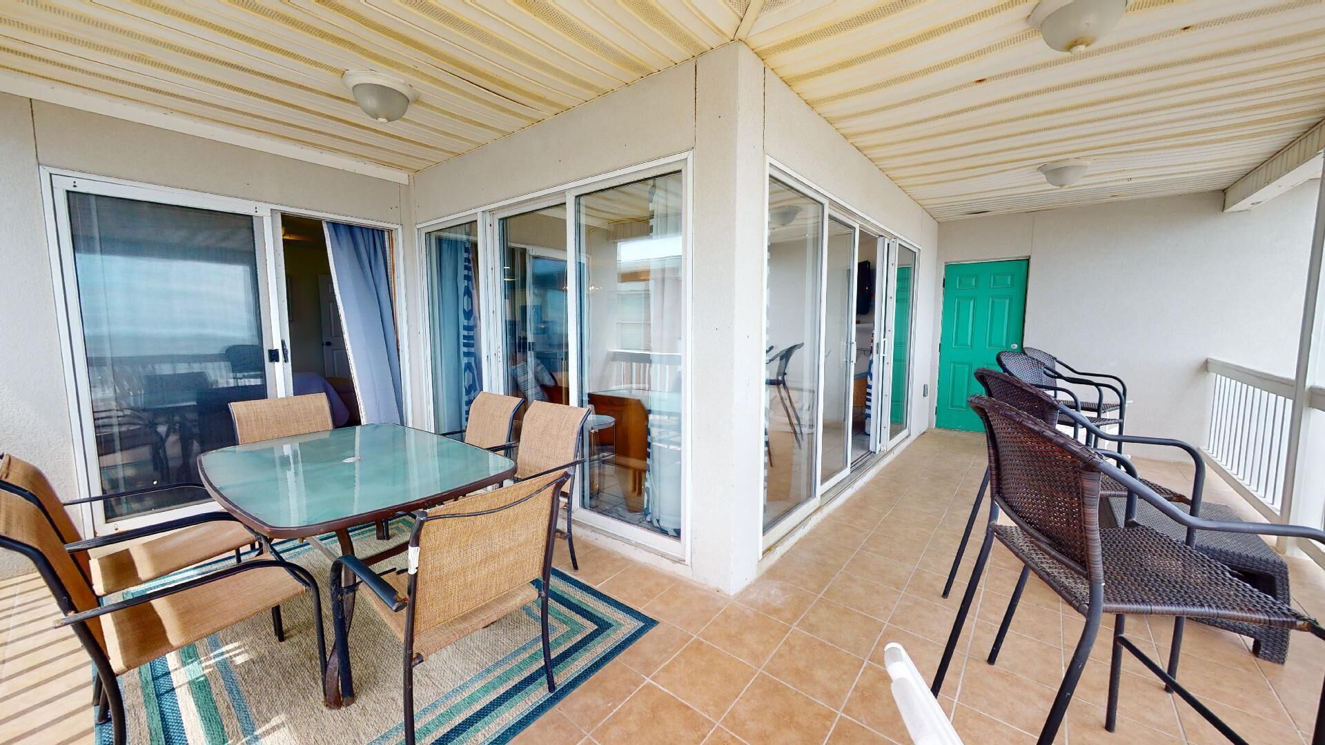 A balcony with a glass table and chairs, a green door, and a view of the sea.