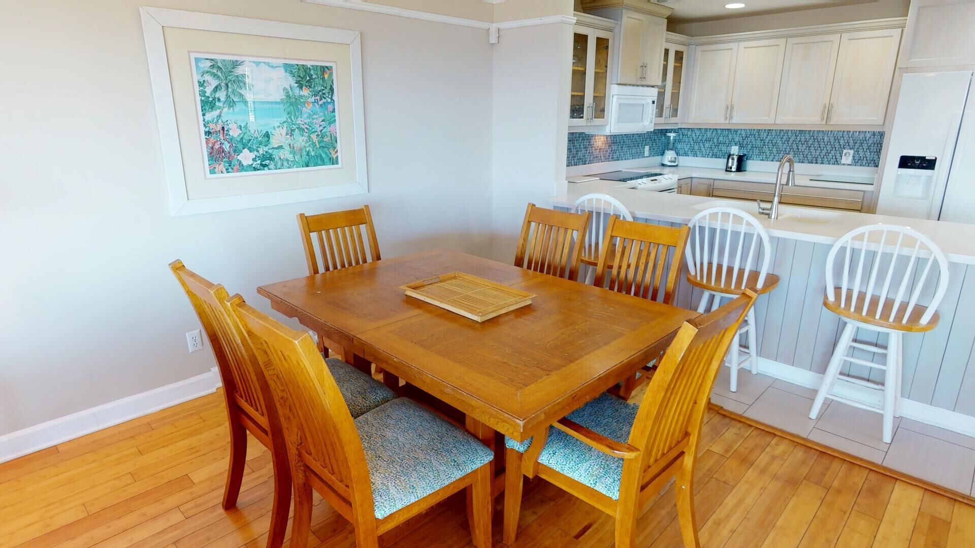 A dining area with a wooden table and chairs, a kitchen in the background, and a framed picture on the wall.