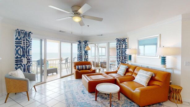 A spacious living room with a large sofa, a coffee table, and a ceiling fan. There is a balcony with patterned curtains and a view of the ocean. - Pelican Point by Tybee Vacation Rentals (Tybee Island)