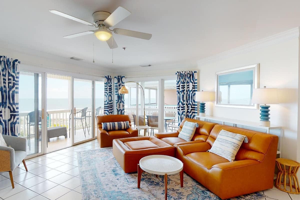 A spacious living room with a large sofa, a coffee table, and a ceiling fan. There is a balcony with patterned curtains and a view of the ocean.