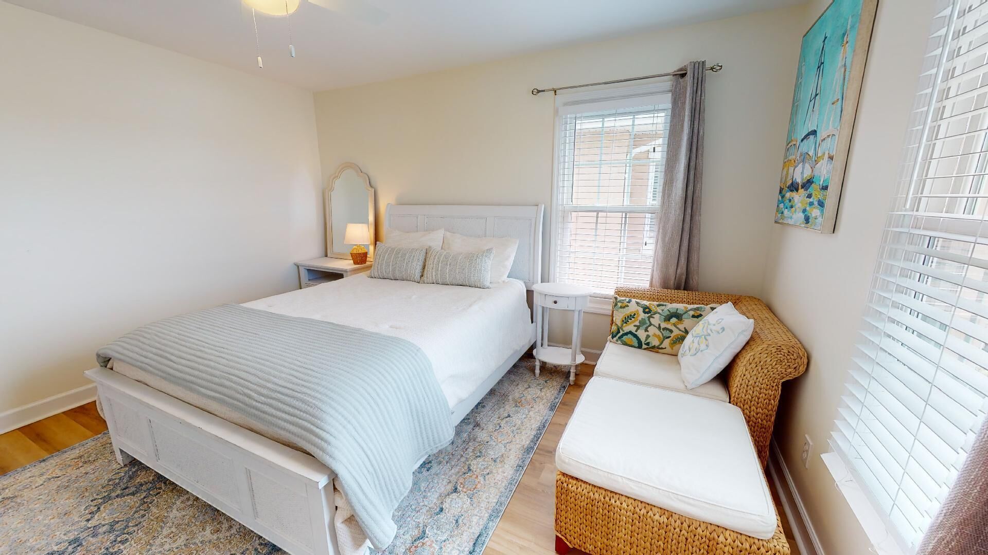 A bedroom with a large bed, a bedside table with a lamp, a small side table, a wicker chair, and a window with blinds.