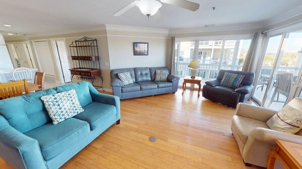 A spacious living room with a blue sofa, a grey sofa, and a beige armchair. There is a ceiling fan, a small table with a lamp, and a picture on the wall. - Pelican Point by Tybee Vacation Rentals (Tybee Island)