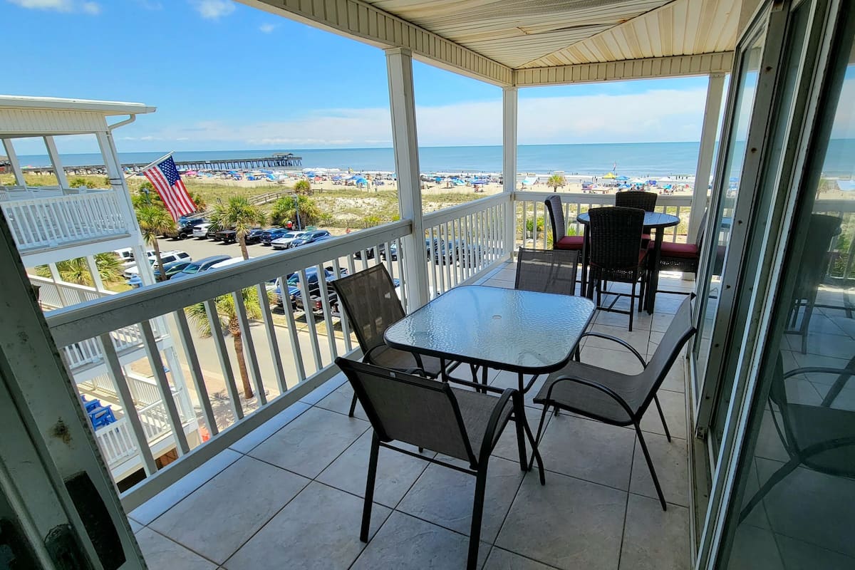 A covered balcony with a glass door, a table, chairs, and a view of the beach and pier.