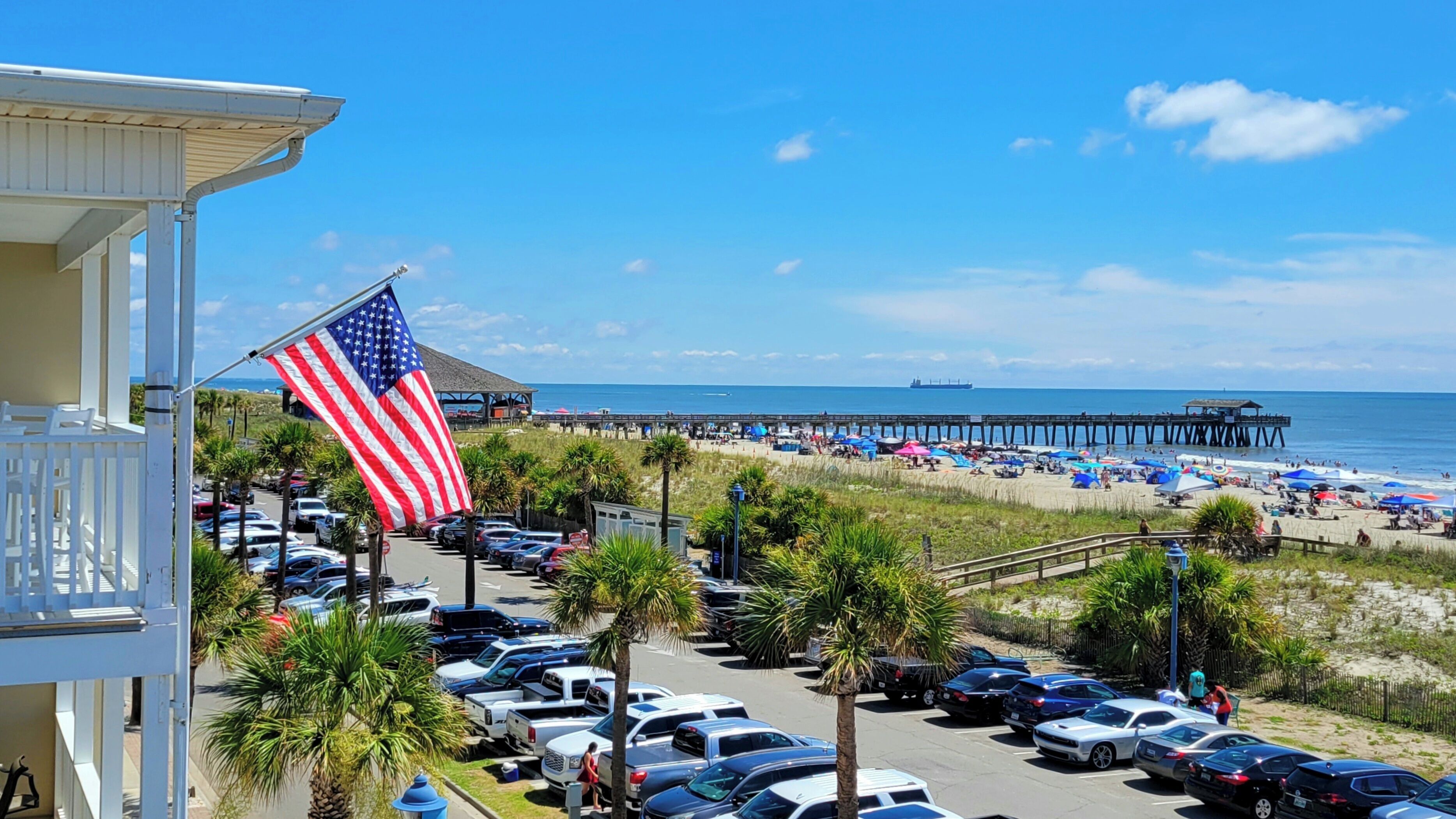 A beachside parking lot with a row of parked cars, a prominent American flag, and a pier extending into the ocean.