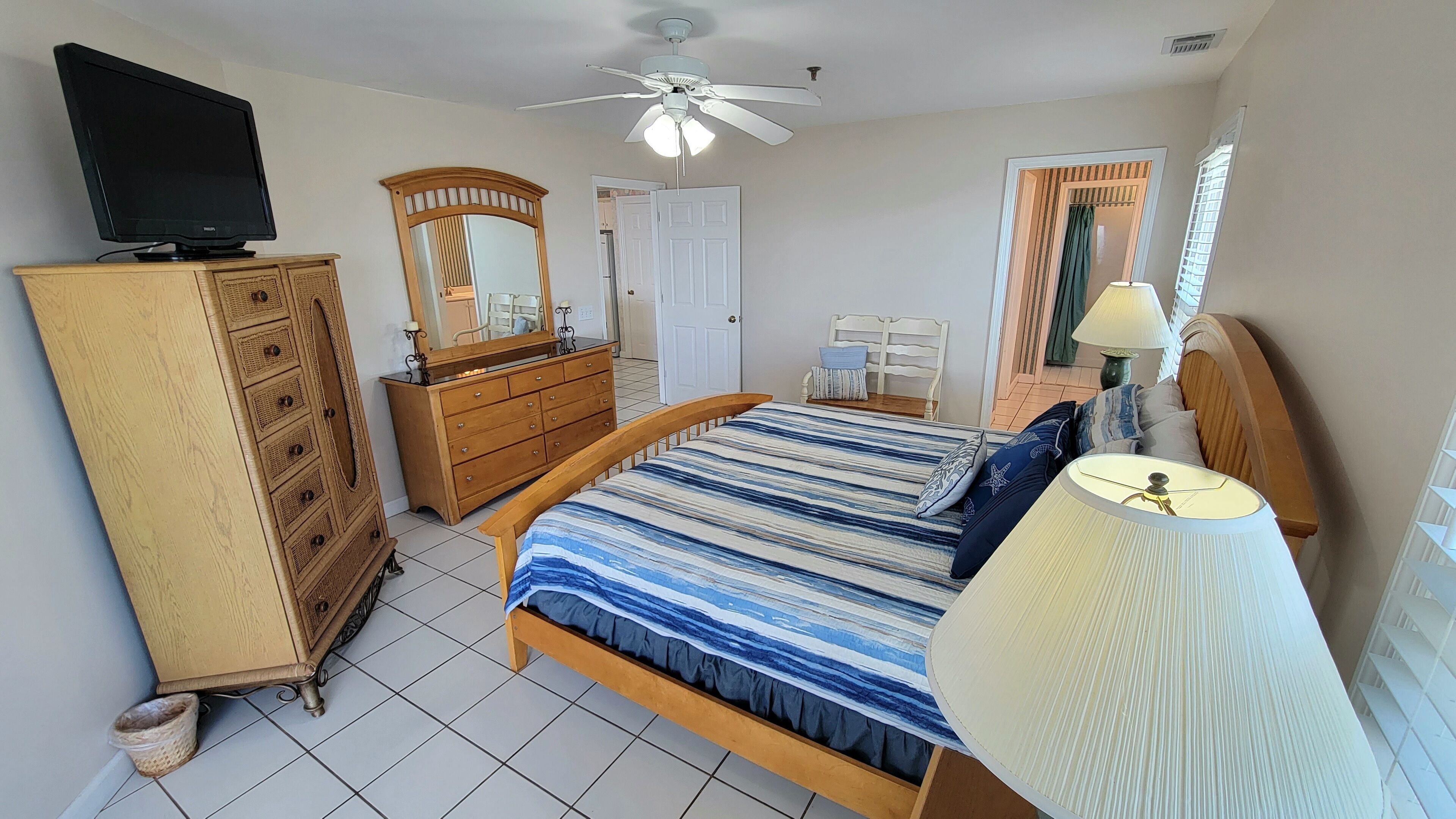 Une chambre à coucher comprenant un lit, une commode, un téléviseur, un ventilateur de plafond et une fenêtre avec des stores.
