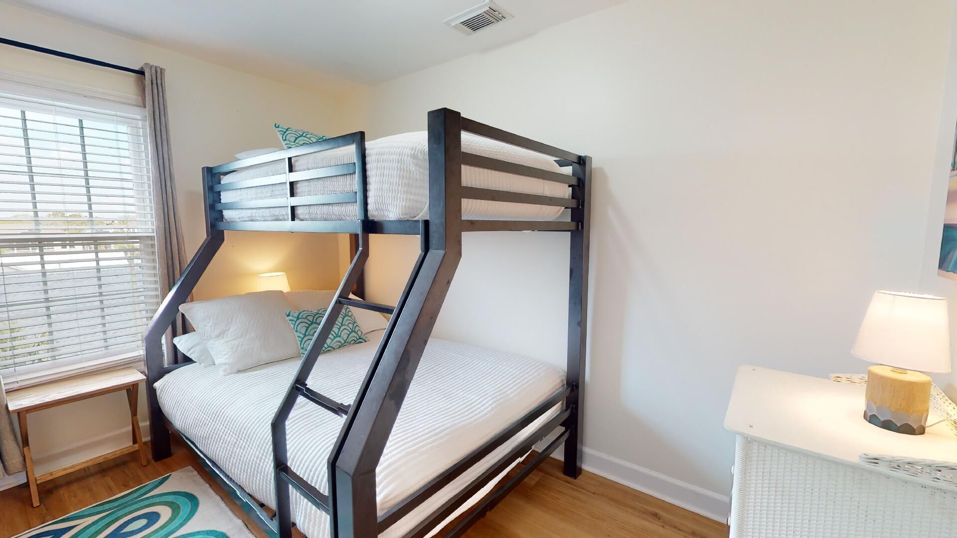 A bunk bed with a desk and lamp, a window with blinds, and a patterned rug.