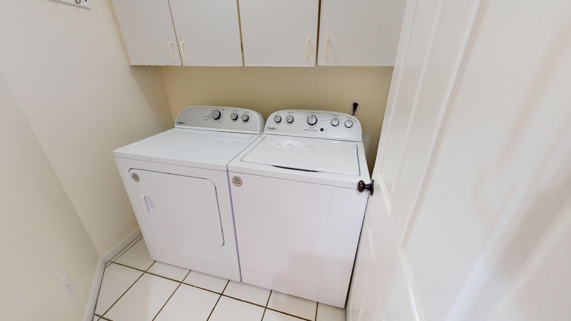 A compact, white, two-door washer and dryer unit in a small laundry room with a tiled floor and white cabinets above.