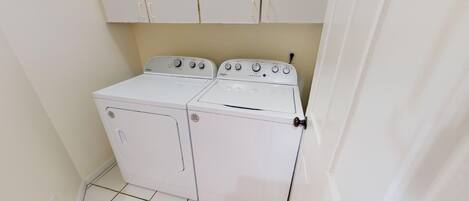A compact, white, two-door washer and dryer unit in a small laundry room with a tiled floor and white cabinets above.