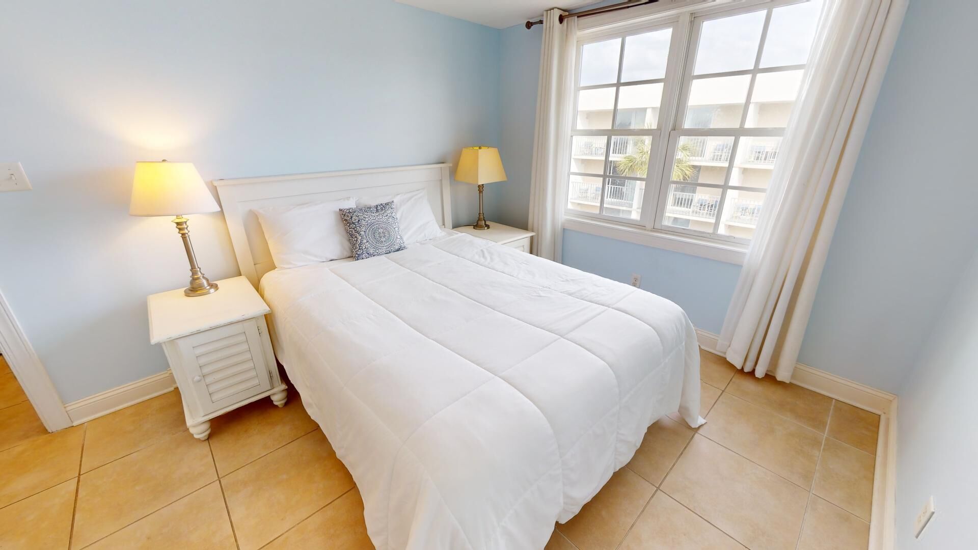 A bedroom with a bed, two bedside tables with lamps, a window with curtains, and a view of buildings outside.