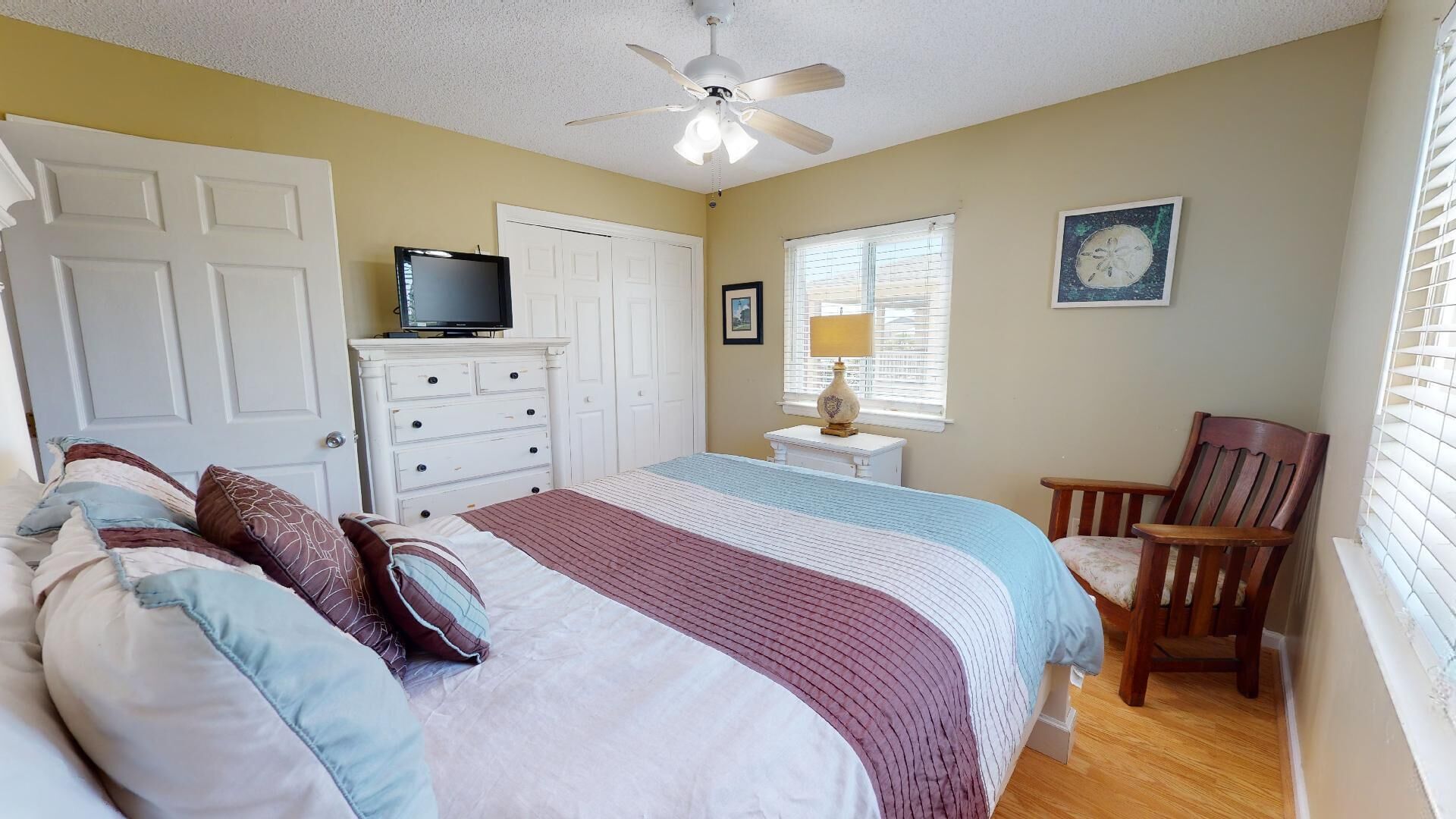 A bedroom with a bed, dresser, television, chair, and a window with blinds.
