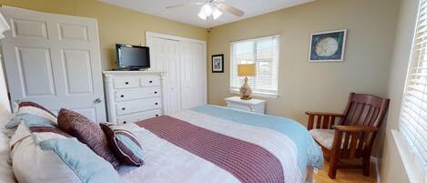 A bedroom with a bed, dresser, television, chair, and a window with blinds.