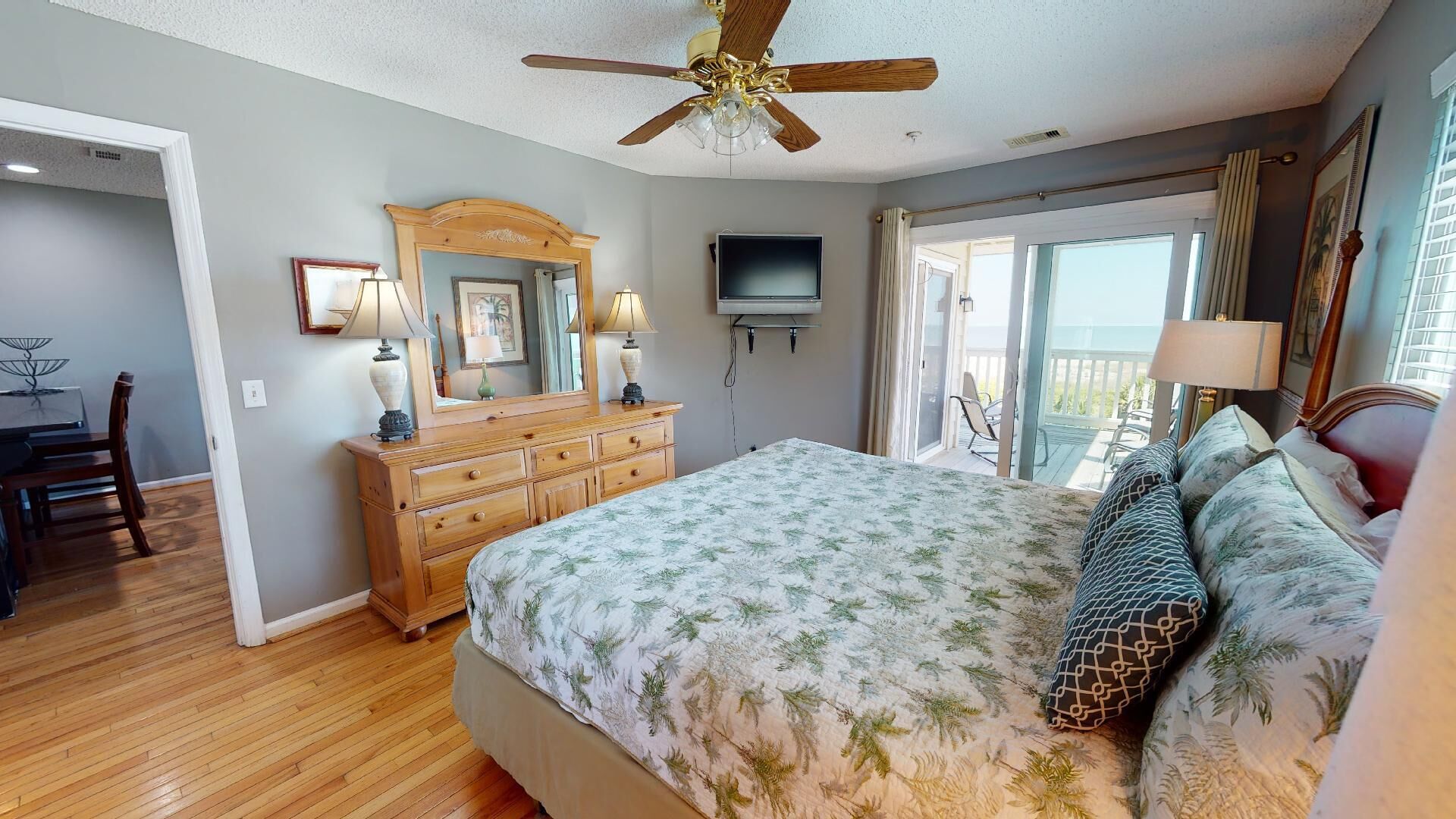 A bedroom with a wooden dresser, a bed with a floral bedspread, a ceiling fan, a television, and a sliding glass door leading to a balcony.