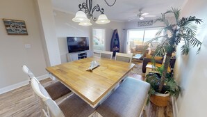 A dining area with a wooden table, white chairs, a TV, and a view of the beach through a large window.