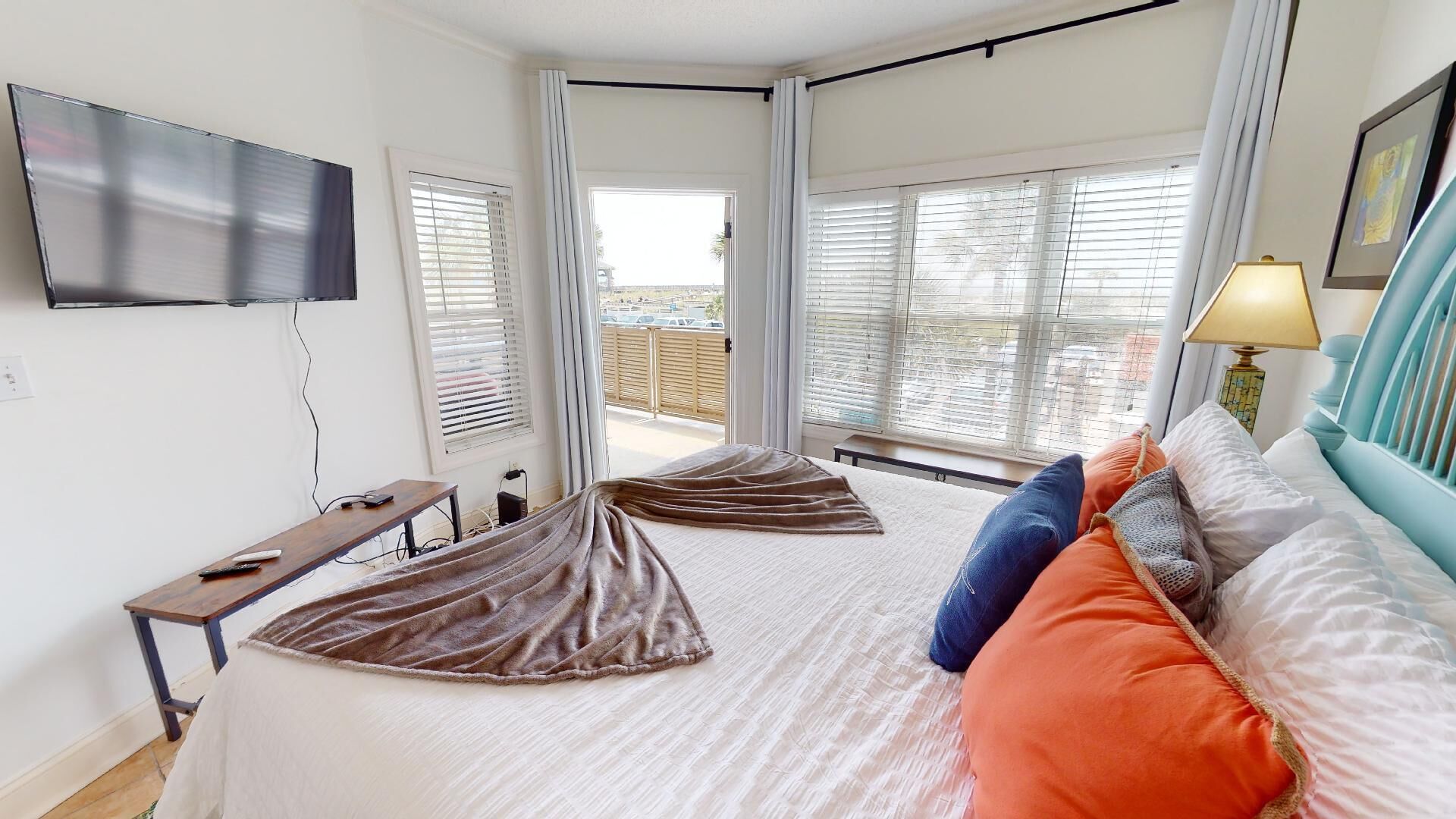 A bedroom with a bed, a TV mounted on the wall, a desk, and a window with blinds.