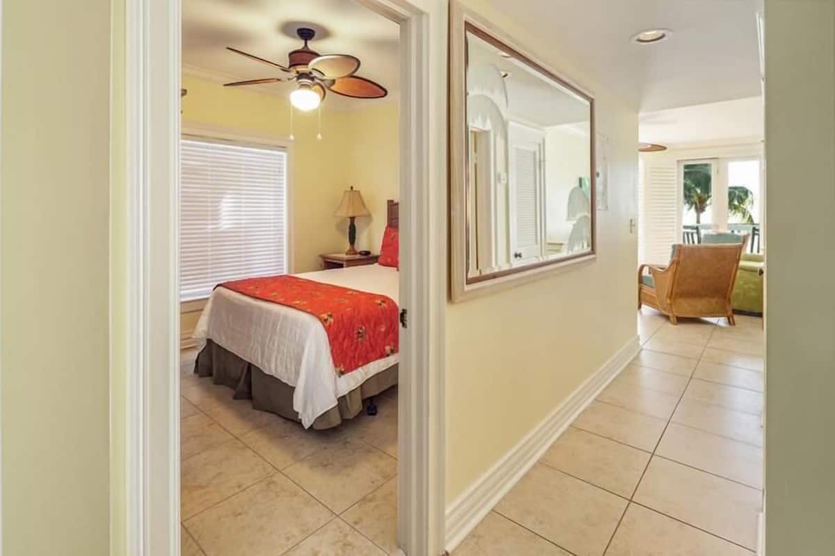 A bedroom with a bed, a ceiling fan, a window with blinds, a nightstand, and a chair in a dining area.